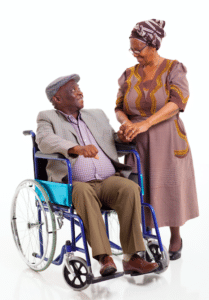 An older man in a wheelchair looks lovingly at an older woman who is holding his hand.