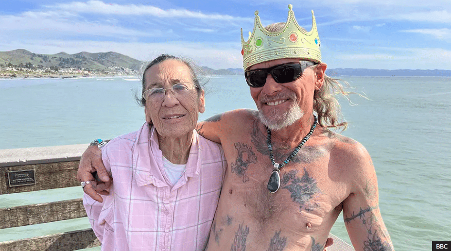 A shirtless man wearing sunglasses and a crown has his arm around an older woman.